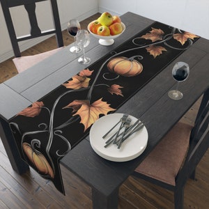 Puede incluir: Un camino de mesa negro con temática otoñal con calabazas naranjas y hojas marrones sobre un fondo negro. El camino de mesa está sobre una mesa de madera oscura con un juego de platos blancos y cubiertos.