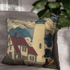 May include: Decorative throw pillow featuring a lighthouse scene. The pillow has a cream-colored lighthouse with a blue top, a white building with a red roof, and a blue sky with white clouds. The pillow is on a gray couch.