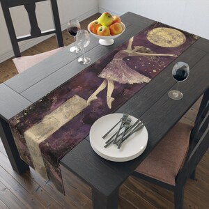 May include: A purple and gold table runner featuring a ballerina dancing in front of a full moon. The runner has a distressed, vintage appearance.