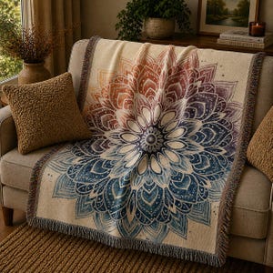 May include: A beige throw blanket with a large, colorful mandala design in shades of blue, purple, orange, and cream. The blanket has fringed edges and is draped over a sofa. A brown textured pillow is on the sofa.