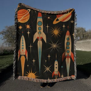 May include: A woven throw blanket with a retro space theme. The design features multiple rockets, planets, and stars in shades of orange, yellow, blue, and red on a black background.