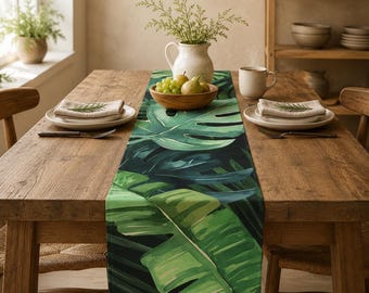 Puede incluir: Un camino de mesa rectangular con un estampado botánico vibrante de hojas grandes y superpuestas de monstera y plátano en varios tonos de verde sobre un fondo oscuro, colocado sobre una mesa de comedor de madera con vajilla.