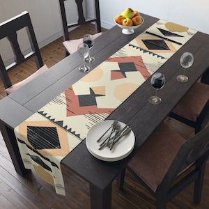 Puede incluir: Un camino de mesa beige con un patrón geométrico en tonos marrones, rosas y negros. El camino de mesa está colocado sobre una mesa de madera oscura con dos sillas.
