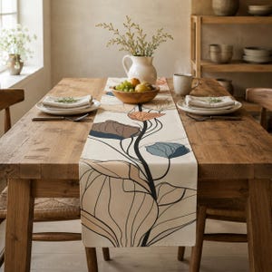 Op de afbeelding: Een crèmekleurige tafelloper met een botanisch lijnpatroon in zwart met accenten in gedempt blauw, oranje en beige. De loper ligt op een houten eettafel die is gedekt met witte borden, gevouwen servetten en een schaal met vers fruit.