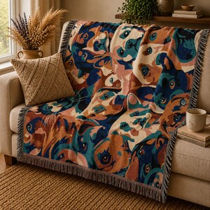May include: A woven throw blanket featuring a repeating pattern of dog faces in shades of blue, brown, and cream. The blanket has a fringed edge and is draped over a beige sofa. A textured pillow and a mug are also visible.