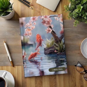 May include: A notebook with a geometric design of two red fish swimming in a pond with pink cherry blossoms above. The notebook cover has the name "CHRISTINA" printed on it.