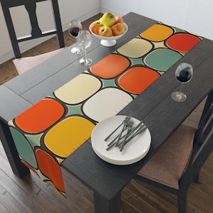 Op de afbeelding: Een tafelkleed met een retro geometrisch patroon van oranje, gele, witte en blauwe cirkels op een teal achtergrond. De loper ligt op een donkerhouten tafel met een bord en bestek.