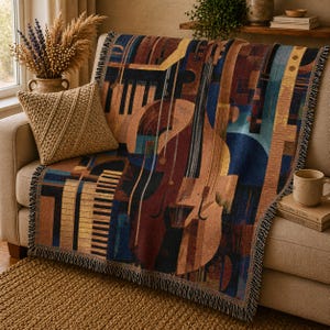 May include: A woven throw blanket featuring a musical instrument design in shades of brown, blue, and yellow. The blanket is draped over a beige sofa, with a matching pillow and a mug visible. The design includes a cello, piano keys, and other musical elements.