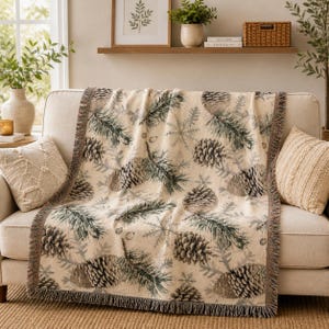 May include: A cream-colored throw blanket with a repeating pattern of pine cones, pine needles, and snowflakes. The blanket has a fringed edge and is draped over a beige sofa. Decorative pillows are on either side of the blanket.