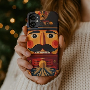 Puede incluir: Una funda de teléfono con una colorida ilustración de un soldado cascanueces. El diseño incluye un sombrero negro, una cara amarilla con una nariz larga y un bigote negro, y un uniforme rojo. La funda se sostiene en una mano.