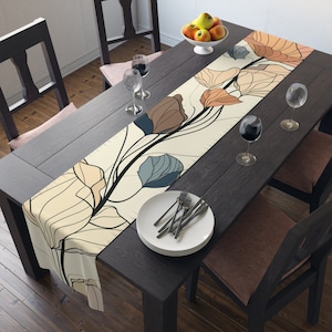 May include: A beige table runner with a floral design featuring brown, blue, and orange leaves. The runner is laid on a dark wood table with two chairs and a set of white plates and silverware.