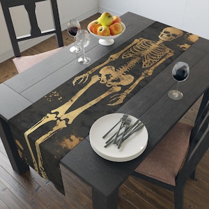 May include: A black and gold skeleton table runner with a distressed look. The runner features a full-length skeleton design with a gold metallic finish.
