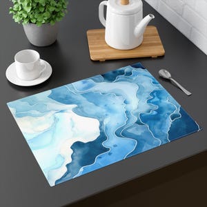 May include: A rectangular placemat with a watercolor design in shades of blue and white. The placemat features flowing, abstract shapes outlined in white, creating a modern and artistic look. The placemat is on a dark surface.