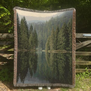 Puede incluir: Una manta tejida con un paisaje de bosque y lago. La imagen muestra el reflejo de los árboles en el agua. La manta tiene un borde con flecos y es principalmente verde, marrón y beige.