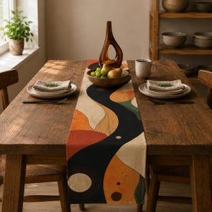 Puede incluir: Un camino de mesa rectangular con un diseño abstracto de formas fluidas en negro, naranja, crema y verde. El camino está extendido sobre una mesa de comedor de madera, decorado con un cuenco de madera con fruta, un jarrón y vajilla.
