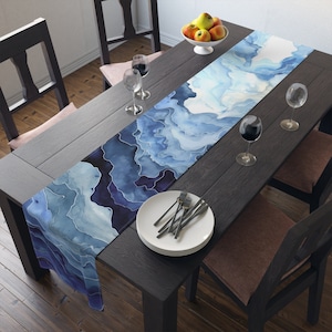 May include: A blue and white watercolor table runner with a swirling abstract design. The runner is laid on a dark wood table with two chairs and glasses of wine.