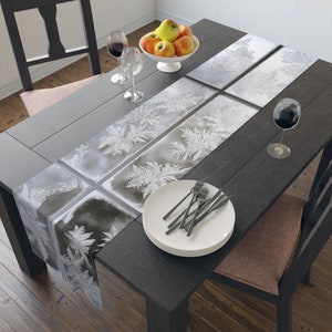 May include: A black and white table runner with a frost pattern. The runner is on a dark wood table with a bowl of apples, two wine glasses, and a plate with silverware.
