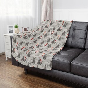 May include: A soft, grey blanket draped over a black leather sofa. The blanket features a repeating pattern of red-hatted figures, grey trees, and bare branches on a light grey background. The blanket adds a touch of festive cheer to the living room.