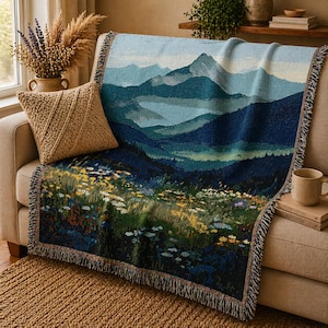 Puede incluir: Una manta tejida con un diseño de paisaje montañoso en tonos azules, verdes y amarillos. La manta está sobre un sofá beige, con un cojín a juego y una taza. La manta tiene un borde con flecos.