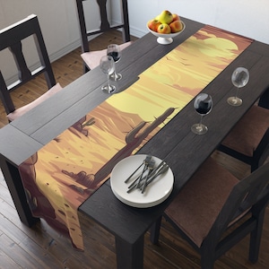 Puede incluir: Un camino de mesa marrón y amarillo con un diseño de paisaje desértico con cactus y una puesta de sol. El camino de mesa está colocado sobre una mesa de madera oscura con dos copas de vino y un plato de cubiertos.