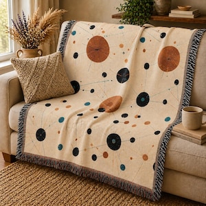May include: Beige throw blanket with a mid-century modern design featuring brown, black, and teal circles and lines. The blanket has a fringed edge and is draped over a sofa. A woven pillow and a mug are also visible.