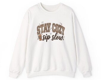 May include: A white crewneck sweatshirt featuring a graphic print on the chest. The text reads STAY COZY in a brown leopard print font and sip slow. in a dark brown script font. A small illustration of a coffee cup with a leopard print sleeve is included.