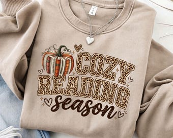 May include: A beige crewneck sweatshirt featuring a graphic of a pumpkin made of stacked books. The text reads COZY READING SEASON in leopard print block letters and brown script. Small hearts and stars surround the design, and a silver heart necklace rests on the collar.