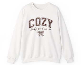 May include: A white long-sleeve crewneck sweatshirt featuring the brown text COZY in a varsity-style font, followed by the cursive script looks good on me and a small brown and white checkered bow graphic centered below the text.
