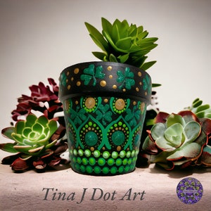 Op de afbeelding: Een handbeschilderde terracotta pot met een groen en gouden klavertjevier-ontwerp. De pot is gevuld met een kleine groene succulent.