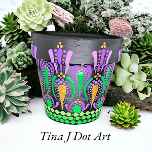 Puede incluir: Una maceta de terracota pintada a mano con fondo negro y un diseño de mandala colorido en tonos de morado, verde y dorado. La maceta está decorada con puntos y espirales. El texto "Tina J Dot Art" es visible en la parte inferior de la imagen.