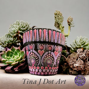 Może przedstawiać: Czarna doniczka z terakoty z namalowanym na niej geometrycznym wzorem w kolorze różowym i fioletowym. Doniczka jest otoczona sukulentami.