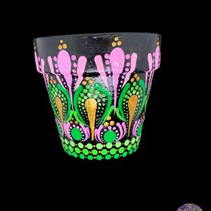 Puede incluir: Una maceta de terracota negra con un diseño pintado a mano. El diseño presenta puntos y remolinos rosas, verdes y dorados. La maceta tiene un diámetro de aproximadamente 10 cm.