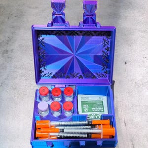 May include: A blue and purple medical kit with a geometric design on the lid. Inside are vials with red caps, syringes with orange plungers, and alcohol prep pads. The kit has two decorative handles.