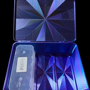 May include: Open, iridescent blue case with geometric starburst pattern on the inside. A clear plastic container with the text "NAD+1" is inside the case. The case has a black background.