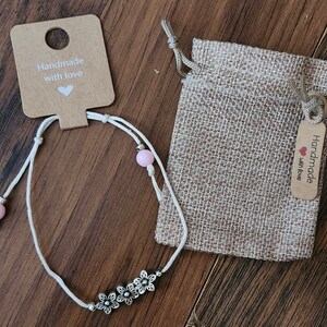 May include: White string anklet with pink beads and silver flower charms. The anklet comes in a small burlap bag with a tag that reads "Handmade with love".