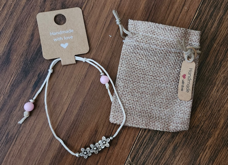 May include: White string anklet with pink beads and silver flower charms. The anklet comes in a small burlap bag with a tag that reads "Handmade with love".