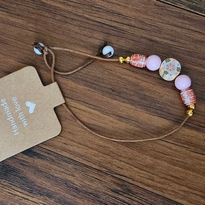 May include: A brown cord necklace with pink and red beads. The beads have a floral pattern and gold accents. The necklace has a tag that says "Handmade with love".