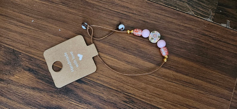 May include: A brown cord necklace with pink and red beads. The beads have a floral pattern and gold accents. The necklace has a tag that says "Handmade with love".