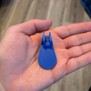 May include: A small, blue, 3D-printed object with a curved, paddle-like base and a raised, rectangular top. The object is held in a hand, and the background is blurred.