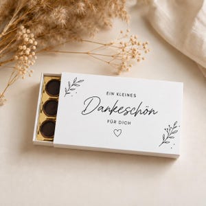 Op de afbeelding: Een witte rechthoekige schuifdoos met drie donkere ronde chocolaatjes in een gouden tray. Het deksel bevat zwarte tekst met de tekst "EIN KLEINES Dankeschön FÜR DICH", een klein hartje en fijne zwarte lijntekeningen van botanische bladeren.