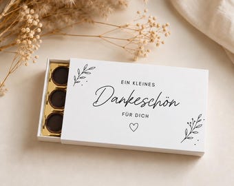 Könnte beinhalten: Eine weiße, rechteckige Schiebeschachtel mit drei dunklen, runden Pralinen in einem goldenen Einsatz. Der Deckel ist mit dem schwarzen Schriftzug "EIN KLEINES Dankeschön FÜR DICH", einem kleinen Herzsymbol und feinen, schwarzen Pflanzenzeichnungen verziert.