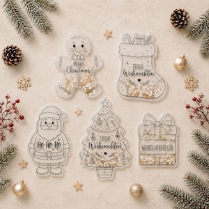 May include: Five clear acrylic Christmas ornaments filled with gold glitter. Shapes include a gingerbread man labeled Merry Christmas, a stocking labeled Frohe Weihnachten, a Santa labeled Ho Ho Ho, a tree labeled Frohe Weihnachten, and a gift labeled Wunscherfüller.