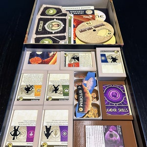 Puede incluir: Un organizador de tablero de juego con compartimentos para cartas, fichas y otras piezas de juego. El organizador está hecho de cartón y tiene un esquema de color negro y dorado. El organizador está etiquetado con los nombres de las diferentes facciones del juego, incluyendo Atreides, Fremen, Tleilaxu y Harkonnen.