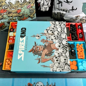 Puede incluir: Juego de mesa "Spires End Rangitaki" con una caja azul claro que presenta una ilustración de guerrero y calaveras. El juego incluye dados y piezas de juego de colores variados en bandejas amarillas, rojas, negras y azules.