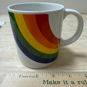 May include: White ceramic mug with a rainbow design in red, orange, yellow, green, and blue. The mug has a curved handle and is on a wooden surface. The text "Make it a rule" is visible on a ruler.