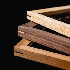 May include: Three rectangular wooden frames stacked at an angle. The top frame is light tan with a visible grain, the middle frame is dark brown with a light inlay, and the bottom frame is medium brown with a dark inlay at the mitered corner joint.