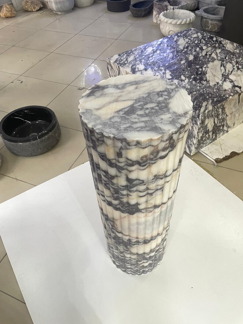 May include: A tall, cylindrical marble pedestal with a fluted design. The stone features a mix of white, gray, and black veining. The top is a flat, circular surface. Other marble pieces are visible in the background.