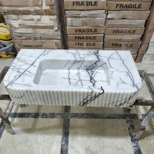 May include: A white marble sink with black veining. The sink is rectangular and has a rounded edge. The sink is sitting on a metal stand.