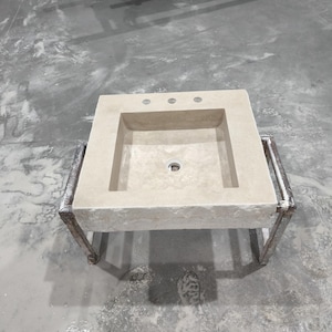 Könnte beinhalten: Ein rechteckiges Waschbecken aus hellem Stein mit einem eingelassenen Becken und drei Wasserhahnlöchern. Das Waschbecken wird von einem Metallrahmen getragen. Das Design ist minimalistisch und für ein Badezimmer geeignet.