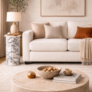 May include: A living room scene featuring a white sofa with decorative pillows in neutral tones. A round marble coffee table holds a bowl of decorative spheres, a small candle, a cup, and books. A marble side table and a framed abstract artwork complete the decor.
